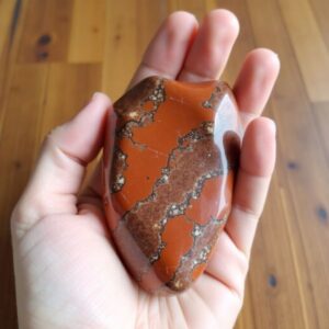 A hand hold a rough red jasper stone against a wooden background